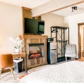 A room with a fireplace, a television mounted above it, a refrigerator, a chair, and a bed.