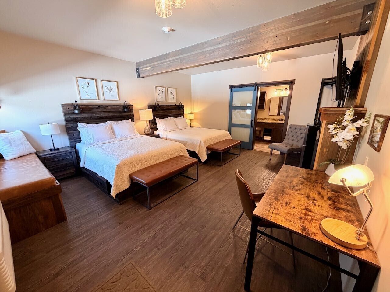 Une chambre d’hôtel avec deux lits, un banc en bois, un bureau avec une lampe et un miroir.
