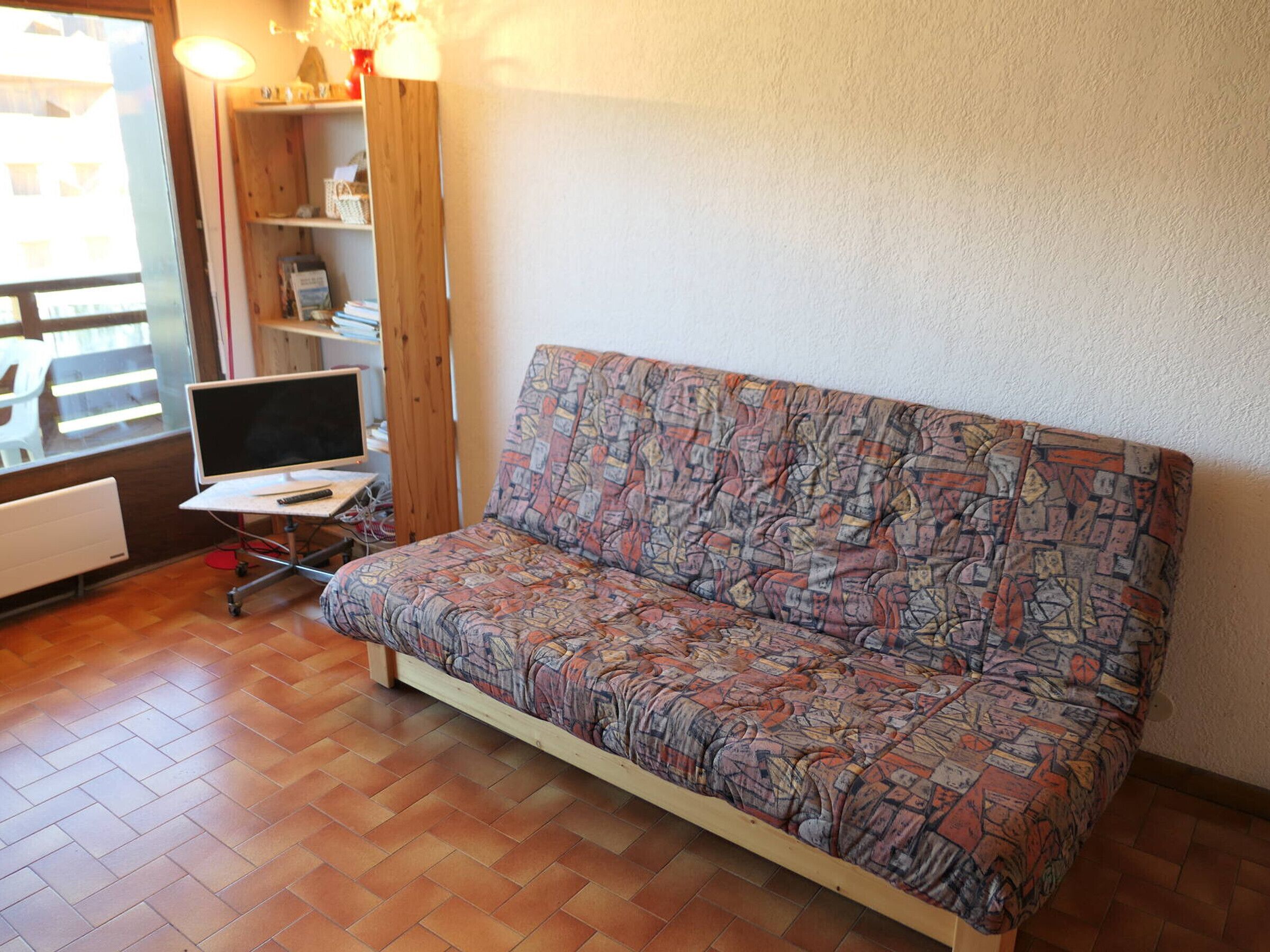 Ein kleiner Raum mit einer gemusterten Couch, einem Schreibtisch mit einem Laptop und einer Heizung.