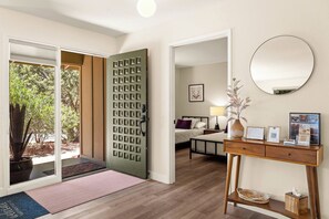A hotel room with a sliding glass door leading to a garden, a bed with a headboard, a bedside table with a lamp, a mirror, and a wooden console table with a vase and a framed certificate.