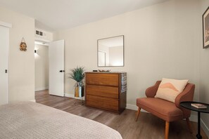 Un dormitorio con cama, cómoda, silla, una mesita y un espejo en la pared.