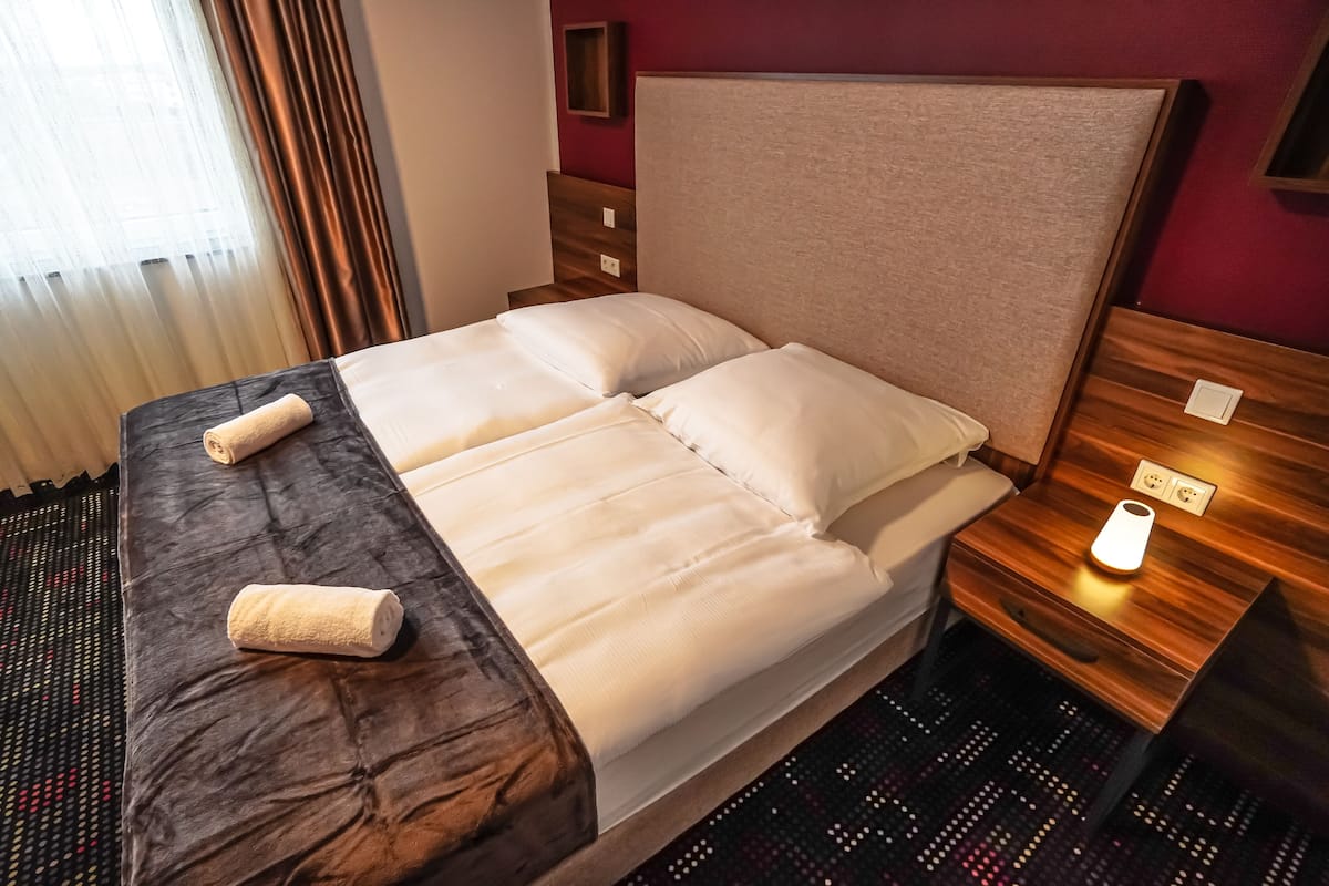 A neatly made bed with a headboard, a bedside table with a lamp, and a window with curtains.
