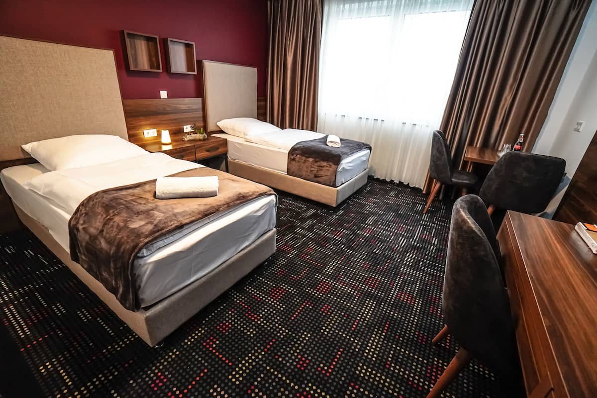 A hotel room with two beds, a desk, a chair, a nightstand, and a window with curtains.