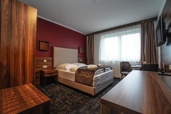 Una habitación de hotel con cama, escritorio, silla, televisión y ventana con cortinas.