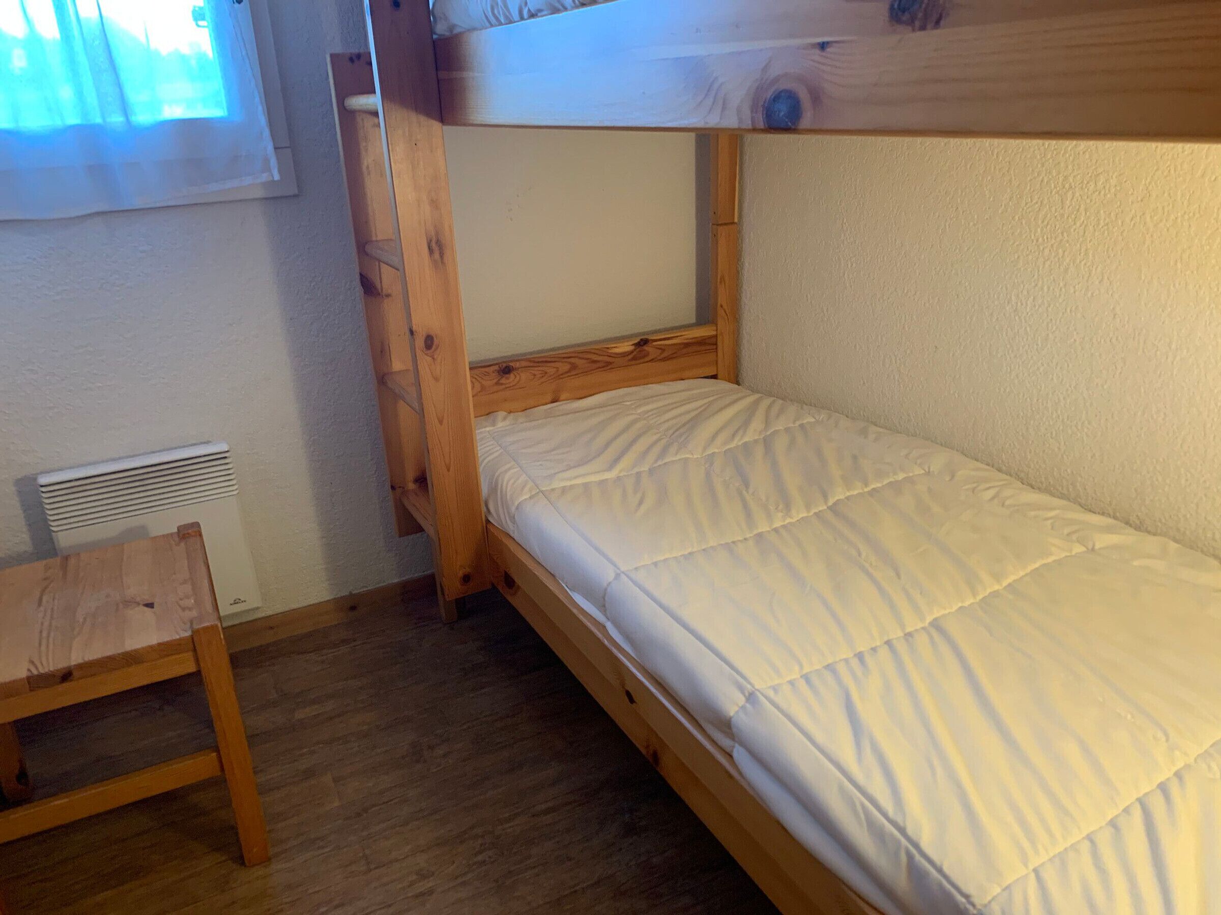 Uma cama beliche com estrutura de madeira, colchão branco e uma pequena mesa de madeira.
