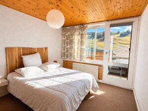 Een slaapkamer met een houten plafond, een groot bed met wit beddengoed, een raam met gordijnen en uitzicht op een landschap.