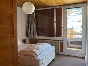 Quarto com cama, radiador, teto de madeira e porta de correr de vidro que dá para uma área externa.