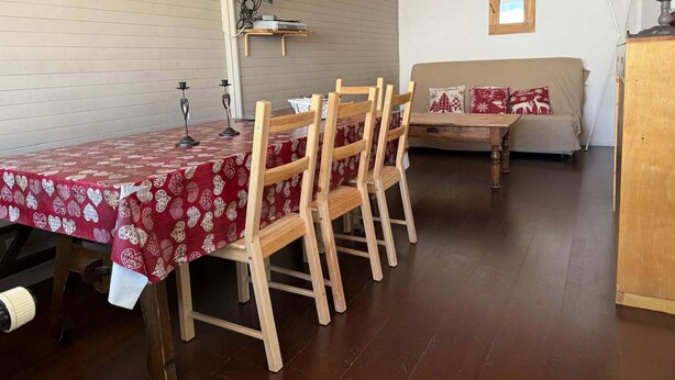 Un coin repas avec une table dressée pour six personnes, des chaises en bois, un téléviseur fixé au mur et un canapé agrémenté de coussins sur le thème de Noël.