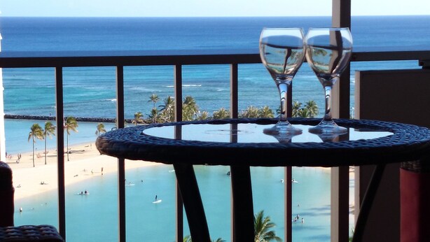 A balcony with a table and two wine glasses, overlooking a beach with people and palm trees. - Posh & Lavish ... 2 ADJOINING SUITES ... Direct Ocean Views (Honolulu)