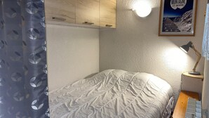 Un dormitorio con cama, escritorio con lámpara, un cartel en la pared y cortina con estampado de hojas.