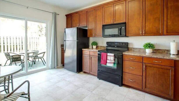 Una cucina con mobili in legno, un frigorifero in acciaio inossidabile, un forno nero e un microonde. C'è un tavolo da pranzo con sedie e una porta scorrevole in vetro che dà su una zona esterna.