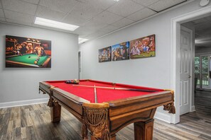 A pool table with a red felt surface, surrounded by wooden legs, in a room with grey walls and a wooden floor.