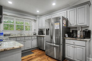 A modern kitchen with white cabinetry, a stainless steel refrigerator, a coffee maker, and a window with blinds.