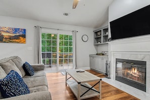 A living room with a sofa, a coffee table, a fireplace, and a large window with a view of greenery.
