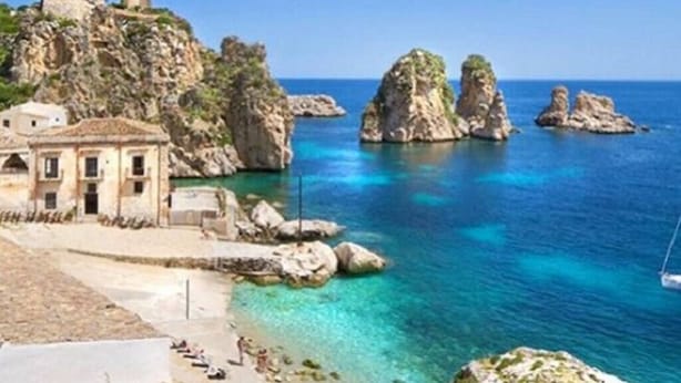 Un edificio sul lungomare con uno sfondo roccioso, acque blu cristalline e una piccola torre.