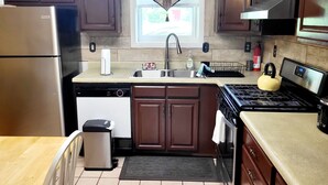 A compact kitchen with dark wooden cabinets, a stainless steel refrigerator, a dishwasher, a sink with a faucet, a stove, and a yellow kettle. - Private and spacious apartment close to Cornell in quiet residential area. (Ithaca)