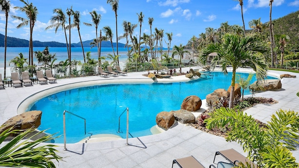 A resort pool with clear blue water, surrounded by palm trees and lounge chairs, with a view of the ocean and mountains in the background. - Whitsunday East 1106 on Hamilton Island (Whitsundays)