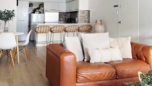 A modern living room with a leather sofa, a dining area with white chairs, and a kitchen in the background. - 2 Spacious Resort 1BR Pool-Tennis-Gym & FREE Parkg (Perth)