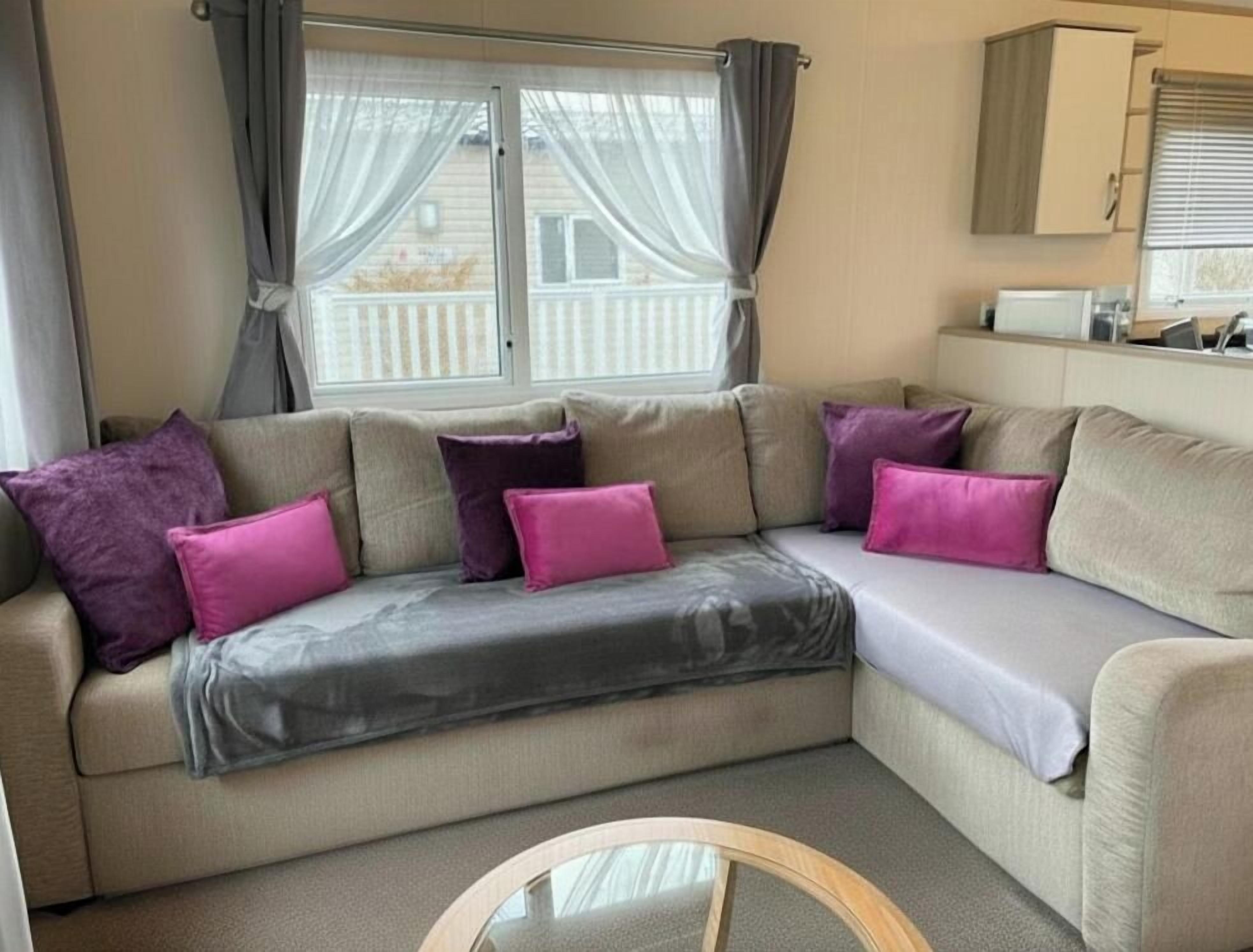 A beige sectional sofa with a grey throw blanket and purple cushions, a round glass coffee table, and a window with curtains.