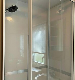 A glass-enclosed shower area with a handheld showerhead, a toilet, and a window with blinds. - Comfortable 3-bed Caravan - Combe Haven (St Leonards-On-Sea)