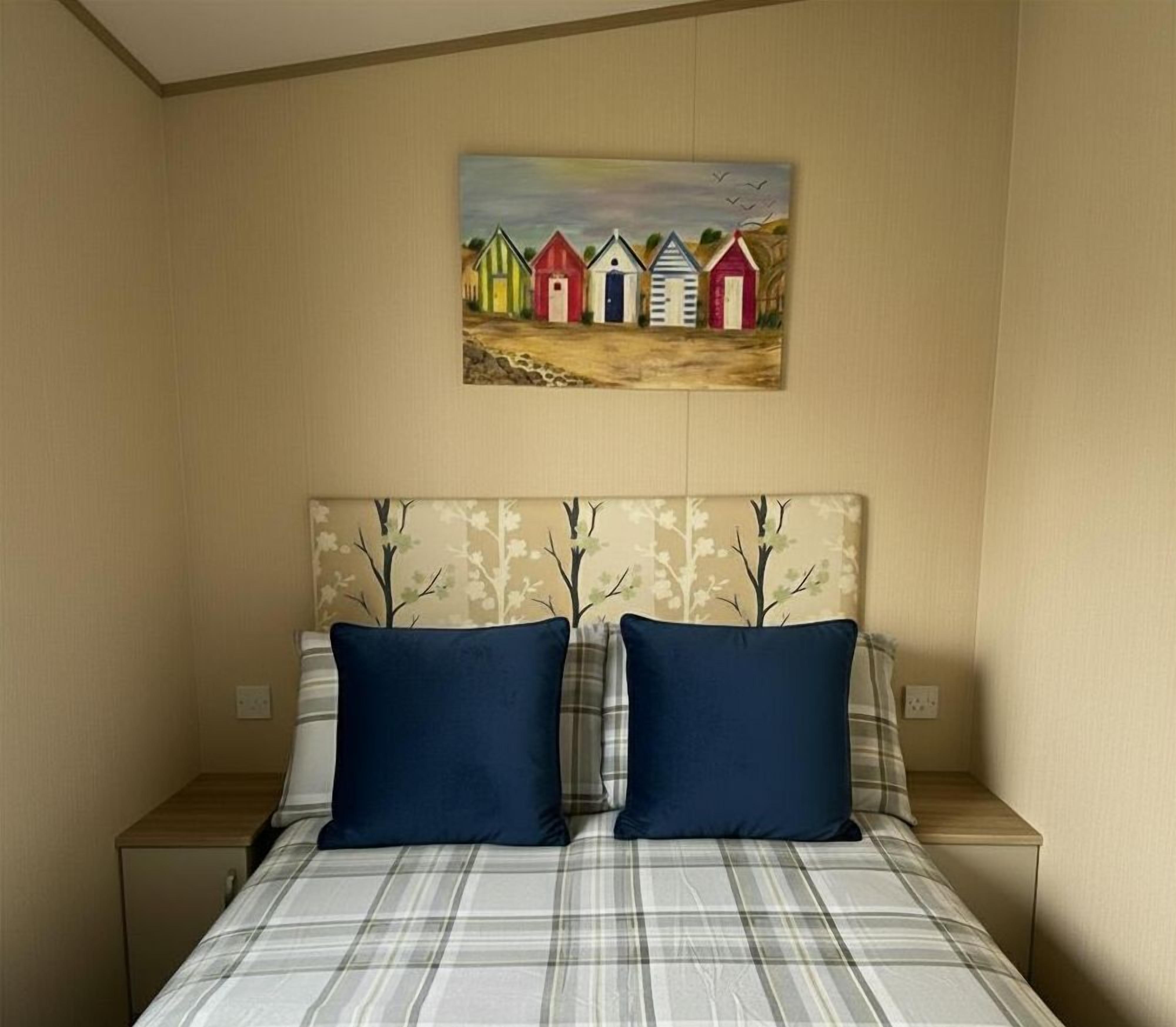 A bedroom with a bed, two pillows, a headboard, a nightstand, and a painting of colorful beach huts.