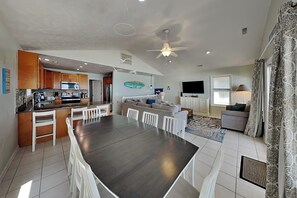 Cozinha moderna com ilha central, sala de jantar com mesa escura e cadeiras brancas, sala de estar com sofá e TV de tela plana, e ventilador de teto.