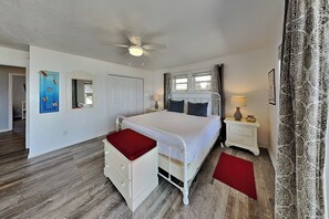 Um quarto com uma cama, uma mesinha de cabeceira, um banco vermelho, um ventilador de teto e uma janela com cortinas.