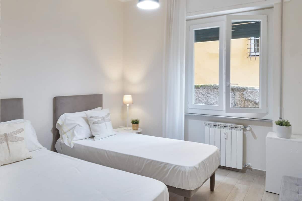 A bedroom with two beds, a bedside table with a lamp, a radiator, and a window with a view of the outside.