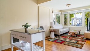 A modern kitchen with a white countertop, a small sink, and a plant. There is a white sofa and a coffee table in the living area, with a view of the outside through large windows. - Fountain Avenue - Pacific Grove Apartment - Steps to the Sea (Pacific Grove)