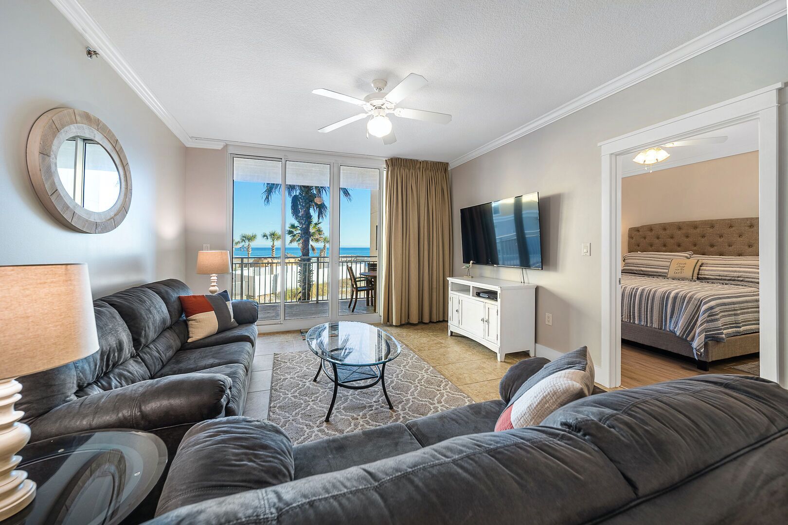A modern living room with a sofa, a glass coffee table, and a TV. There is a balcony with a view of palm trees and the ocean.
