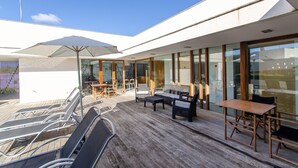 A modern outdoor lounge area with sun loungers, a wooden deck, and glass doors leading to an interior space. - Villa “Ses Alzines” with Sea View, Pool, A/C, Wi-Fi, Balcony, Garden & Terrace (Sant Lluís)