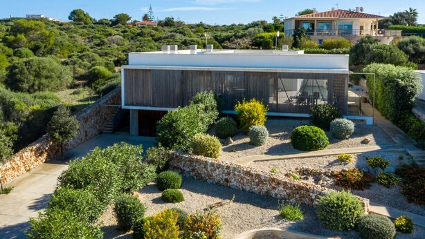 A modern house with a flat roof, surrounded by a well-maintained garden with various shrubs and a stone wall. - Villa “Ses Alzines” with Sea View, Pool, A/C, Wi-Fi, Balcony, Garden & Terrace (Sant Lluís)
