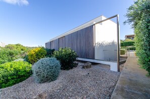 A modern building with a sloped roof, surrounded by gravel and various shrubs. - Villa “Ses Alzines” with Sea View, Pool, A/C, Wi-Fi, Balcony, Garden & Terrace (Sant Lluís)