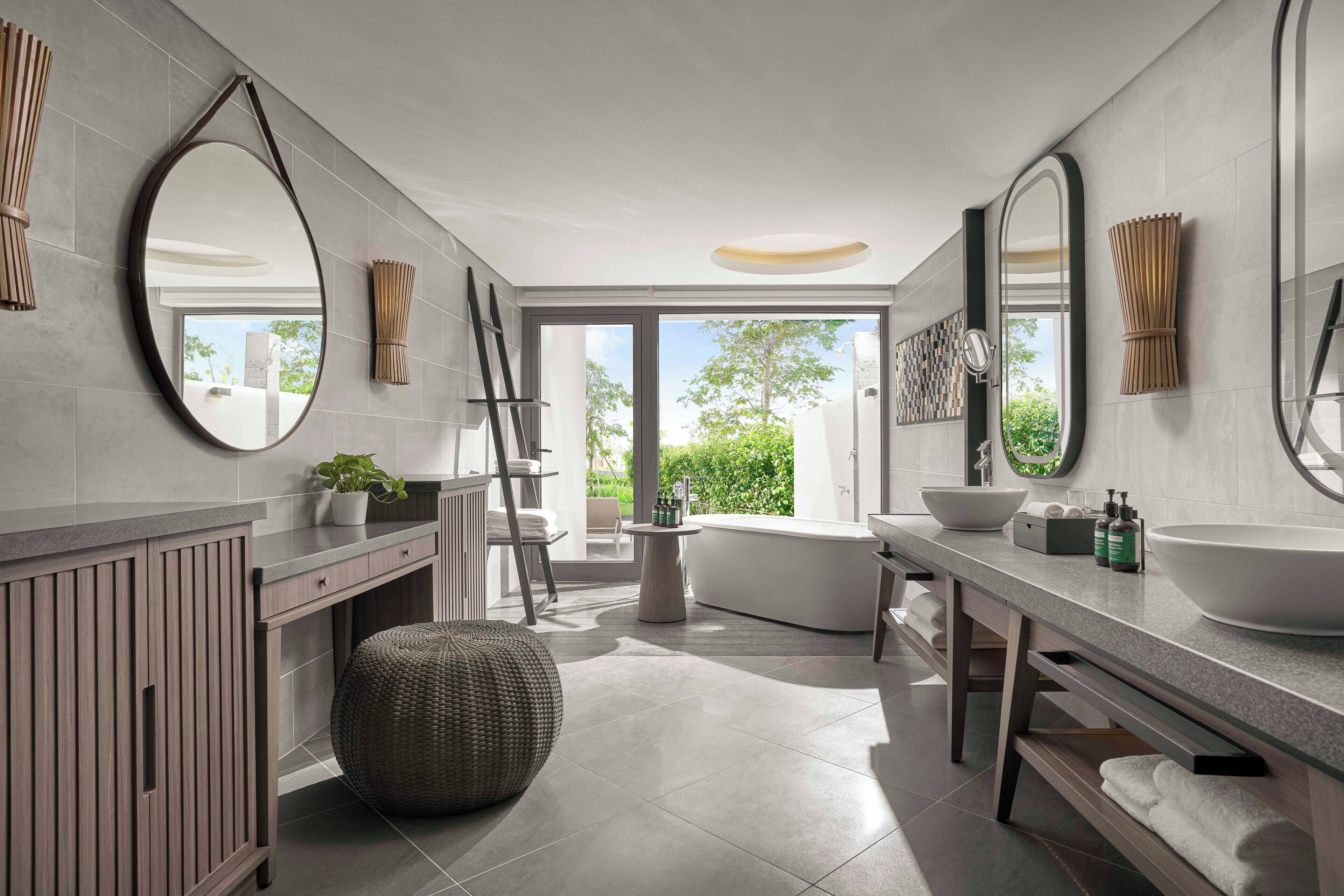 A modern bathroom with a large mirror, two sinks, a bathtub, and a large window providing a view of greenery outside.