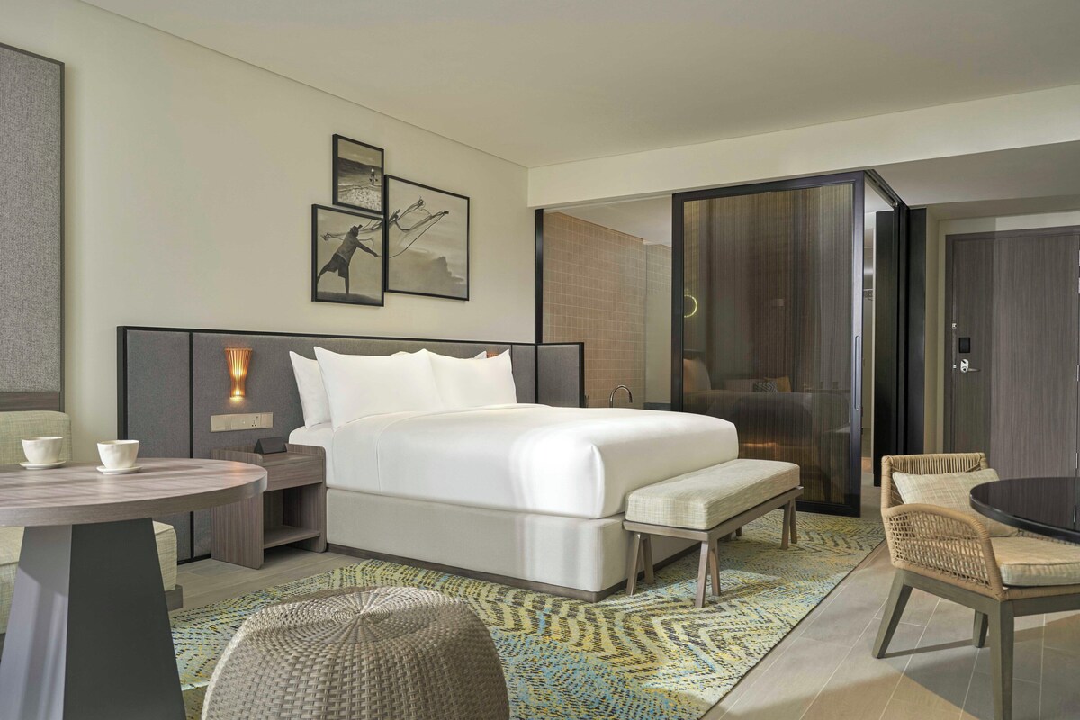 Een moderne hotelkamer met een groot bed, een kleine tafel, een stoel en een bank. Er hangen afbeeldingen in lijsten aan de muur en er is een raam met uitzicht.