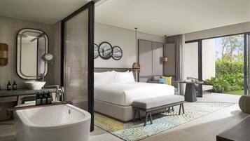A modern hotel room with a large bed, a bathroom with a bathtub, a sitting area with a table and chairs, and a view of greenery outside.