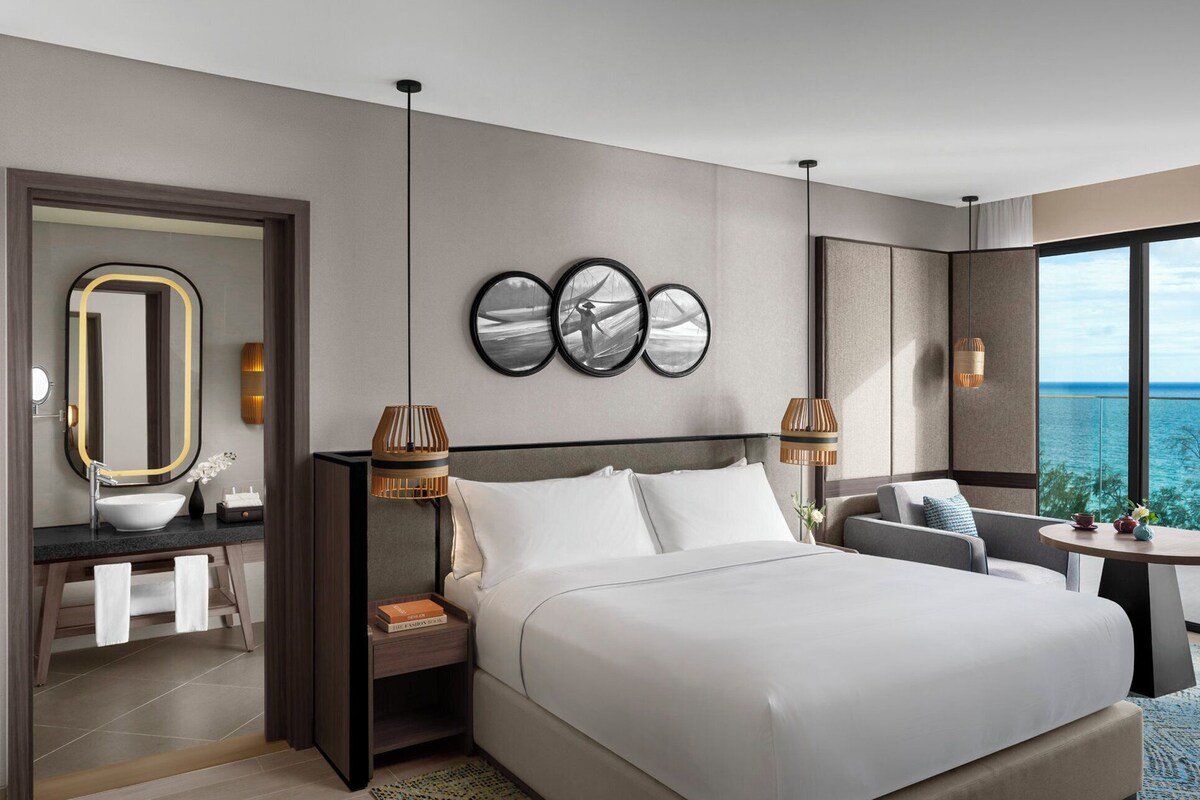 Een moderne hotelkamer met een groot bed, een kaptafel met spiegel en uitzicht op de oceaan.