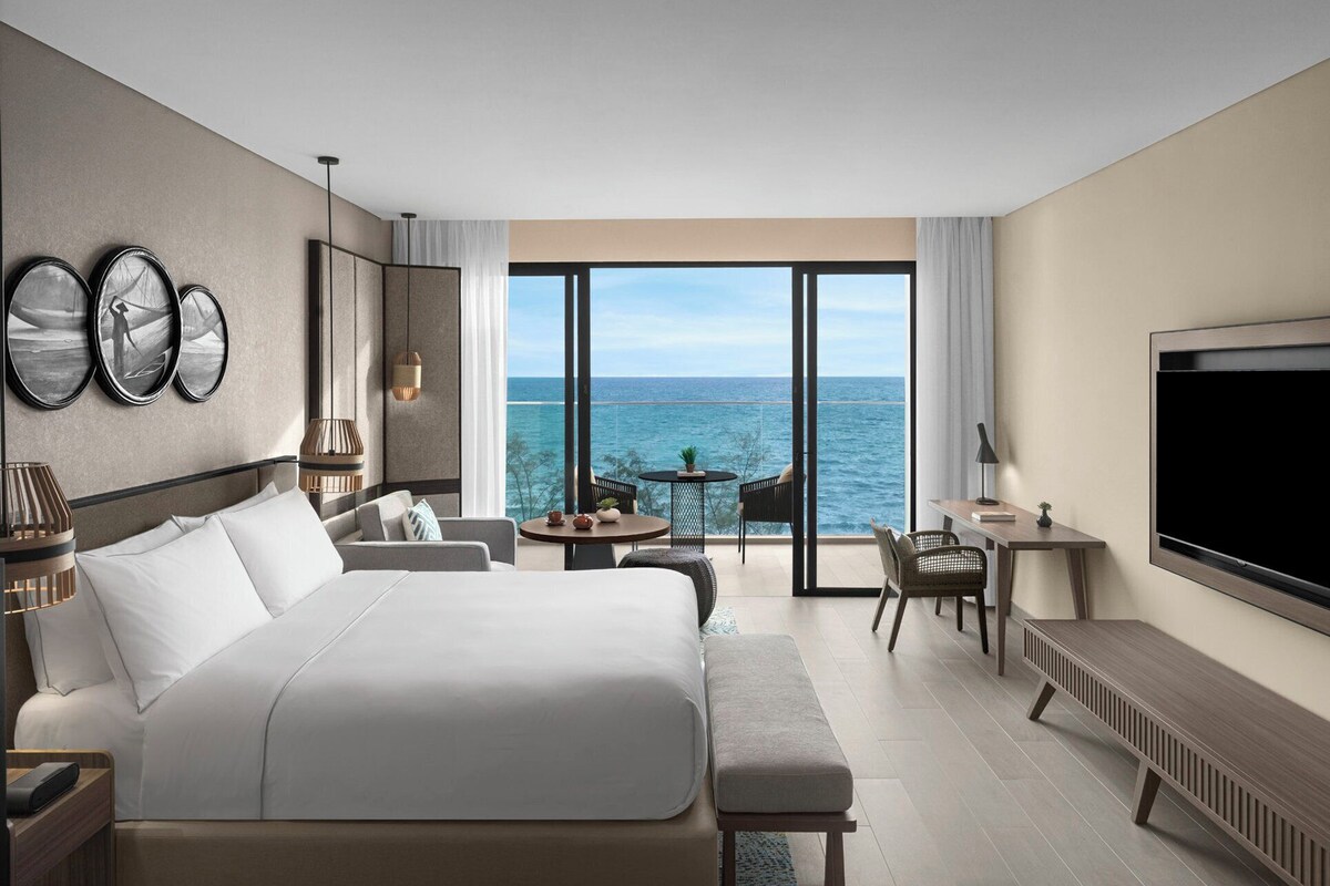 Une chambre d’hôtel moderne dotée d’un grand lit, d’une télévision et d’un coin repas avec une table et des chaises, et offrant une vue sur la mer.
