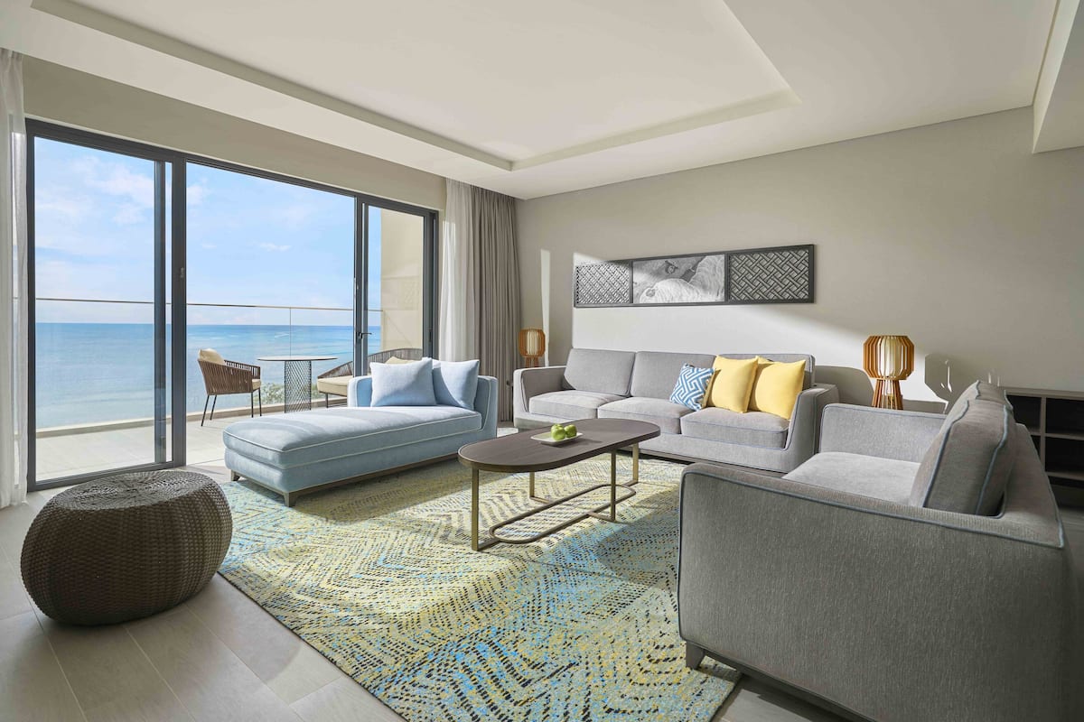 Een moderne woonkamer met een grote schuifpui die leidt naar een balkon met uitzicht op de oceaan. Er staan twee banken, een salontafel en een kamerkleed met een patroon.