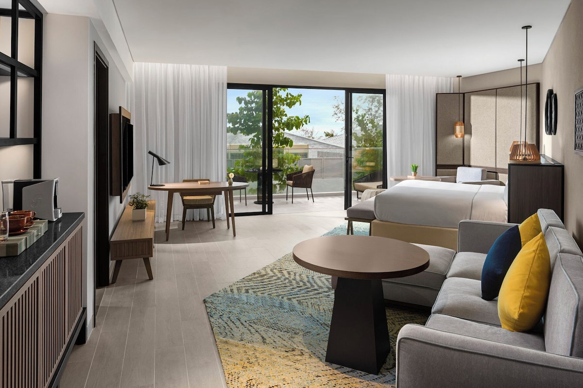 Een moderne hotelkamer met een groot bed, een bank, een eettafel en een televisie. Er is een balkon met uitzicht op de omgeving.
