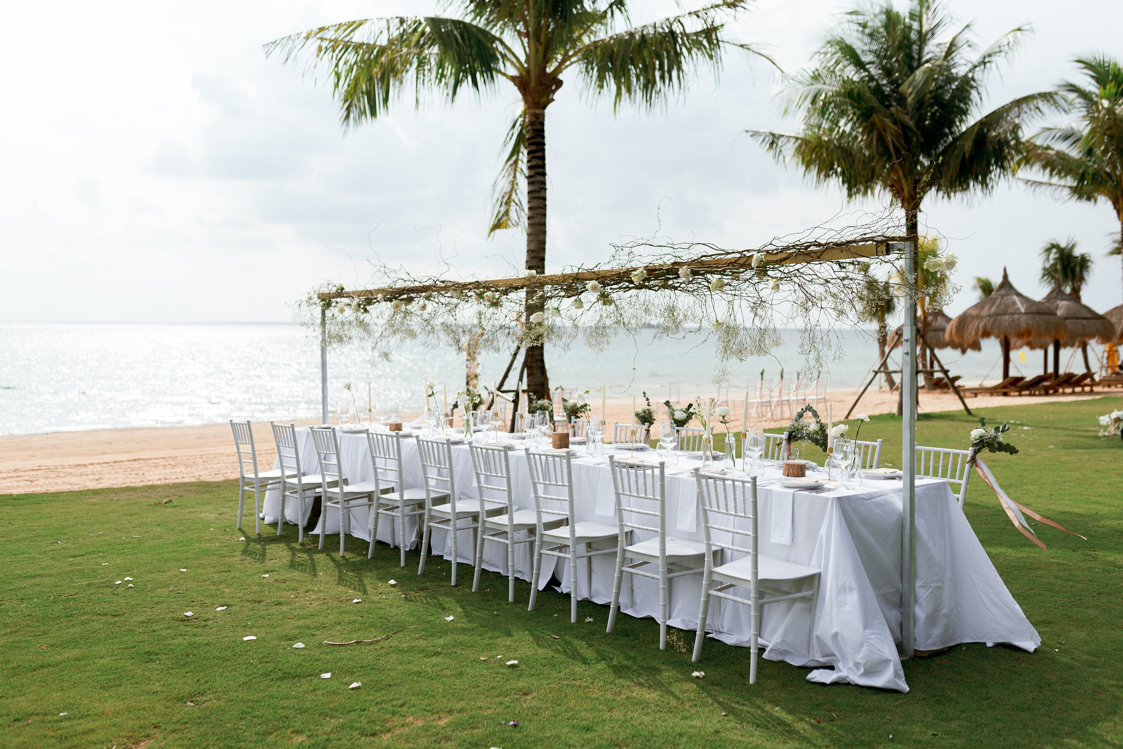 A beachside wedding setup with white tables and chairs, palm trees, and a thatched structure in the background.