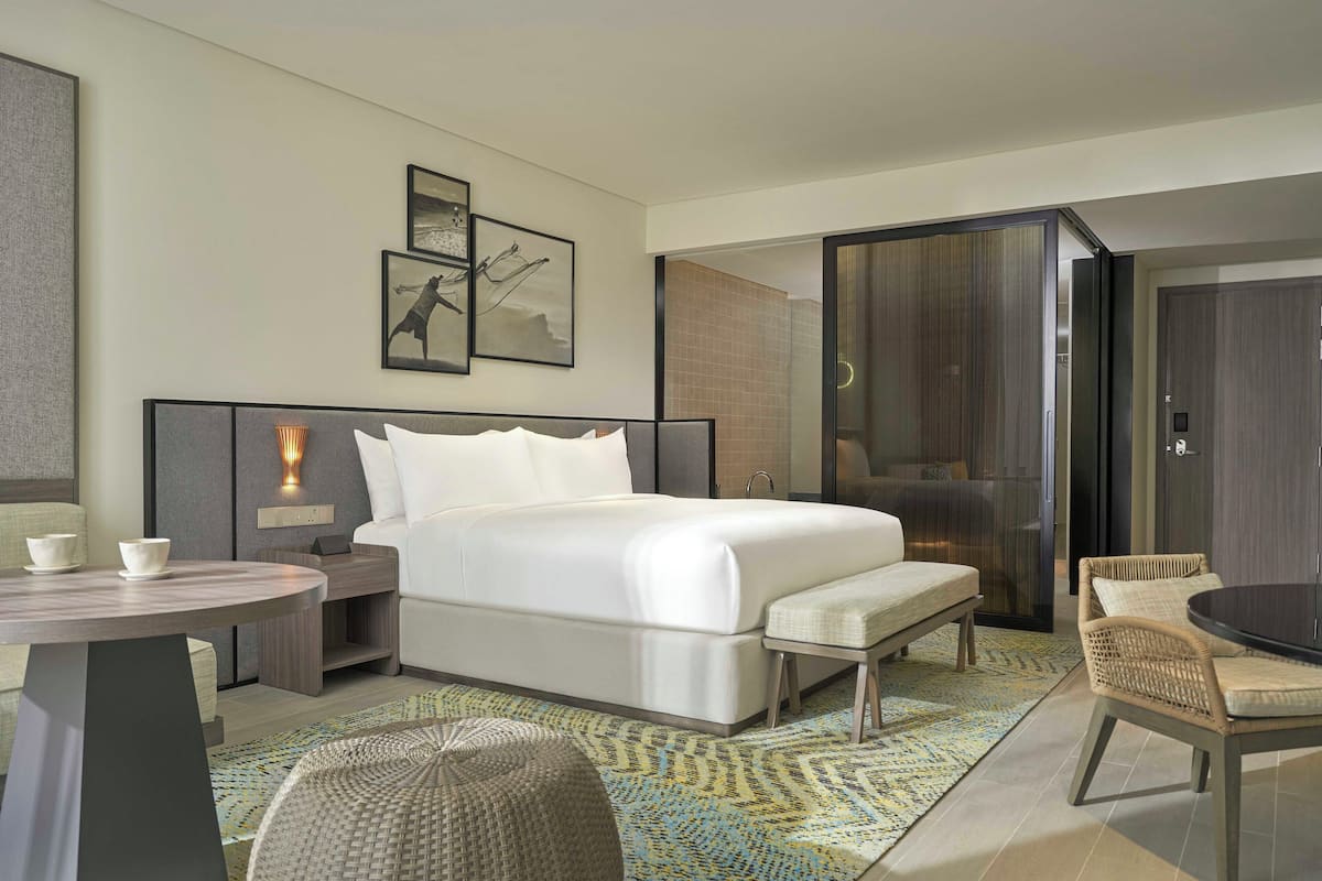 Een moderne hotelkamer met een groot bed, een kleine tafel, een stoel en een bank. Er hangen afbeeldingen in lijsten aan de muur en er is een raam met uitzicht.