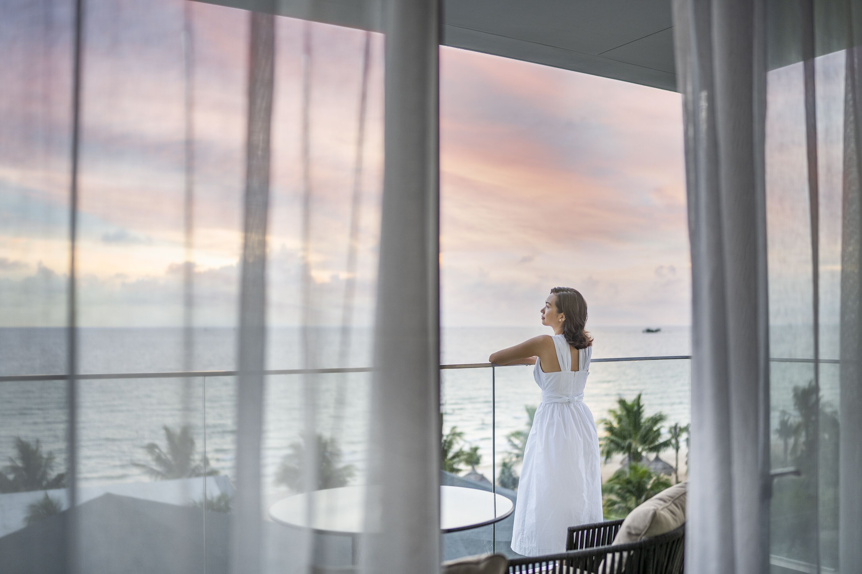 A woman in a white dress standing by a balcony with a view of the ocean and palm trees.
