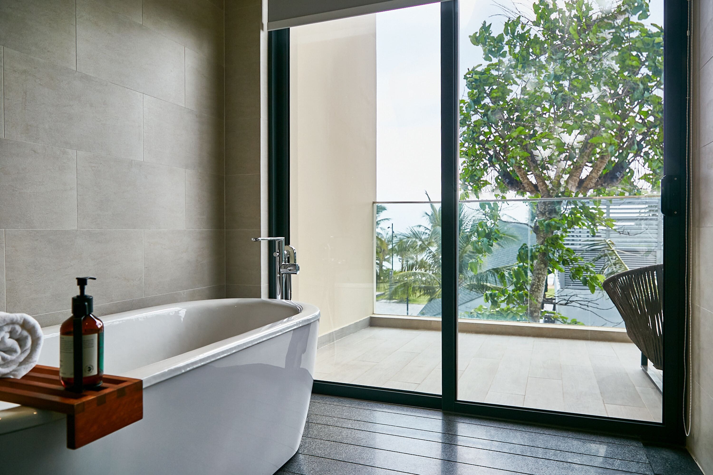 A modern bathroom with a freestanding bathtub, a glass door leading to a balcony with a view of greenery and a tree.
