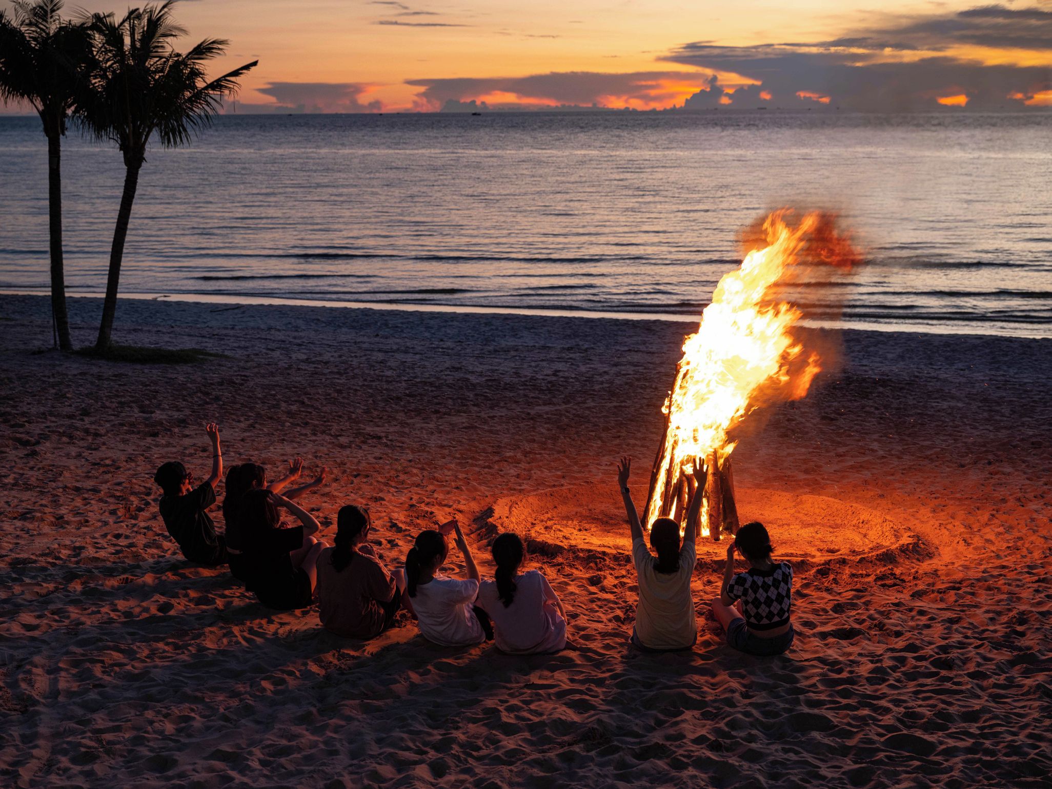 A beach scene with a fire in the sand, palm trees, and the ocean in the background.