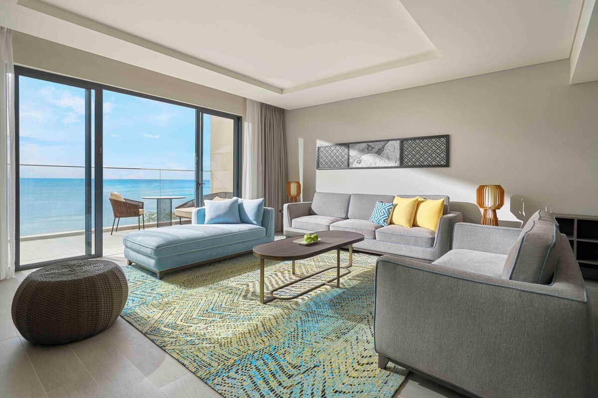 Een moderne woonkamer met een grote schuifpui die leidt naar een balkon met uitzicht op de oceaan. Er staan twee banken, een salontafel en een kamerkleed met een patroon.