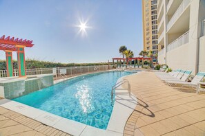 A swimming pool with a sunshade structure, lounge chairs, and a tall building in the background.