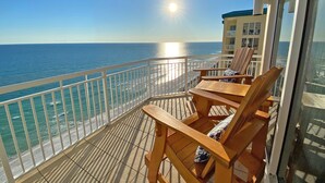 Un balcon avec des chaises et une table en bois, donnant sur une plage, avec l’océan en vue et un bâtiment en arrière-plan.