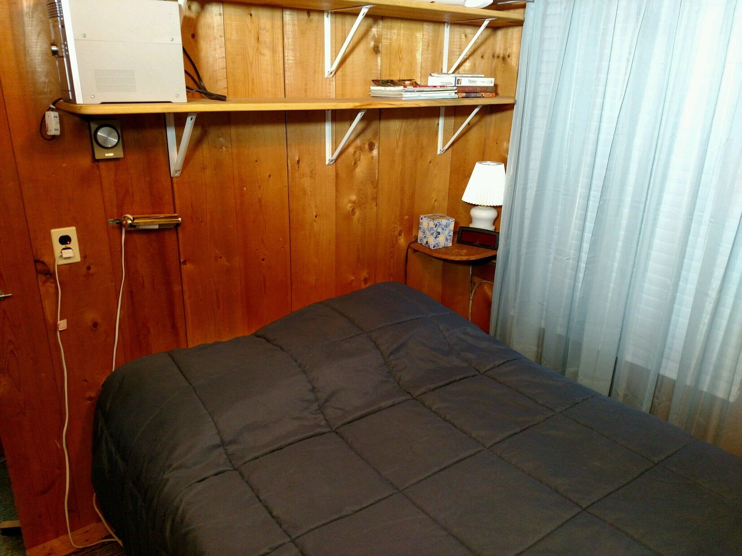 A room with a bed, wooden walls, a shelf with books, a lamp, and a power outlet.