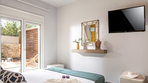 Une chambre à coucher avec un lit, un téléviseur fixé au mur, une fenêtre donnant sur l’extérieur et une petite table sur laquelle se trouvent un vase et un chapeau.